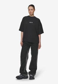 Zwarte oversized t-shirt met korte mouwen en een vierkant logo op de borst, gecombineerd met donkerdenim wijde broek en chunky zwarte sneakers.