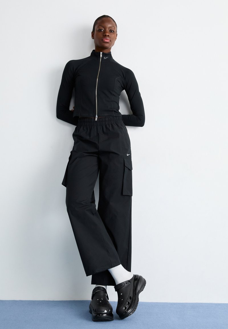 Schwarze Zip-up-Jacke mit hohem Kragen, schwarze Cargo-Hosen mit elastischem Bund und schwarze Clogs. Das Modell trägt weiße Socken.