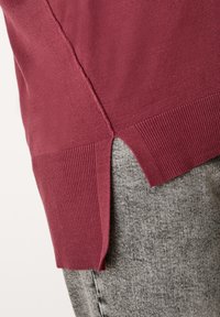 Gros plan de l'ourlet latéral d'un pull en maille rouge avec un détail de fente porté sur un pantalon gris texturé.