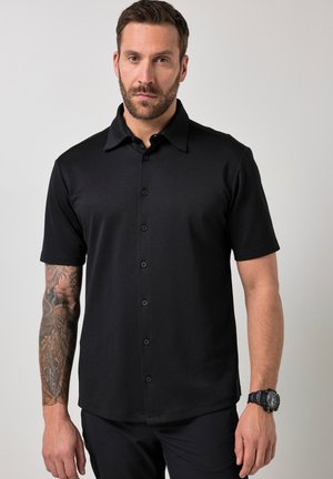 Man met baard die een zwart kortermig button-up overhemd draagt, tatoeage op de linkerarm en een zwart polshorloge aan de rechterpols tegen een lichte achtergrond.