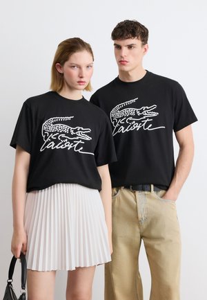 Zwei junge Erwachsene, die schwarze Lacoste-T-Shirts mit weißem Krokodil-Logo tragen; Frau in weißem Faltenrock hält eine schwarze Handtasche; Mann in beigen Hosen.