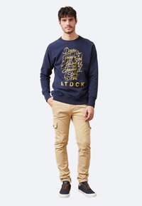 Sudadera azul marino con diseño de texto amarillo, pantalones cargo color beige y zapatillas azul marino. Ajuste casual con puños y dobladillo acanalados.