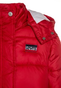 Rød dunjakke med høj krave, hvide skulderdetaljer og et fremtrædende broderet Levi's-logo på brystet. Tekstureret stof med lodrette sømme.