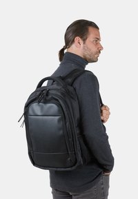 Mochila negra hecha de un tejido duradero, con un panel frontal sintético liso, cremalleras y correas acolchadas. Bolsillo lateral de malla visible.