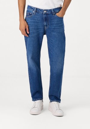 Mann trägt blaue Jeans mit geradem Bein und weiße Sneakers, steht mit einer Hand in der Tasche vor einem einfarbigen Hintergrund.