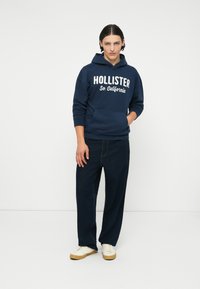 Mornarsko moder hoodie z belim napisom "HOLLISTER So. California", kengurska žep; v kombinaciji s temno jeans hlačami in kremnimi supergami.