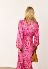 Ξανθιά γυναίκα με μακρύ ροζ φόρεμα με floral σχέδιο, κρατώντας καφέ σουέντ τσάντα, στέκεται μπροστά σε μια απλή ανοιχτή τοίχο.