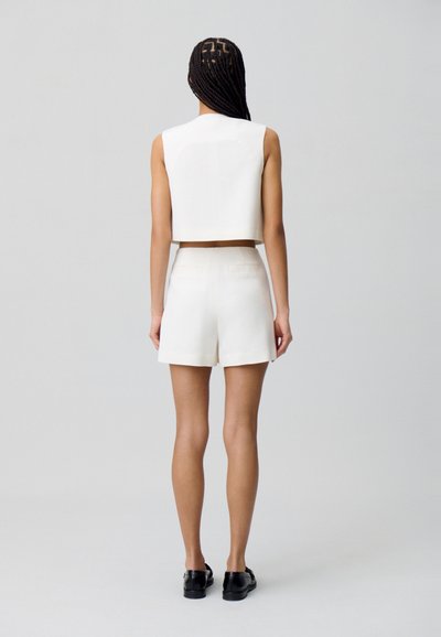 Haut court blanc et shorts taille haute, avec une texture lisse, un design minimaliste et des poches arrière. Des chaussures plates noires complètent le look.