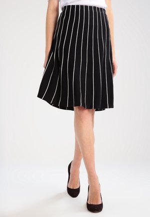 Vrouw draagt een zwarte rok met witte verticale strepen en zwarte hoge hakken, staand met gekruiste benen tegen een witte achtergrond.