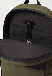 Grønn nylonryggsekk med svart fôr, et merke, elastiske stropper og en QR-kode. Har glidelås og rund toppdesign.