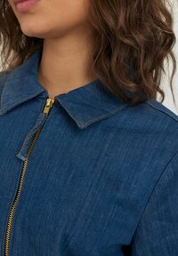 Chaqueta de denim de color azul oscuro, con cuello puntiagudo, cremallera dorada y detalle de costura visible. El material tiene una textura suave.