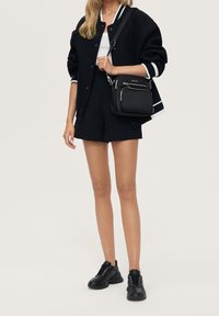 Černá univerzitní bunda s bílým lemováním, bílý top, černé šortky a černé tenisky. Taška má několik kapes na zip a elegantní design.