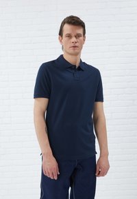 Mornarska polo majica, kratki rukavi, klasični ovratnik s dva dugmeta, napravljena od glatkog materijala. Nosiva s tamnoplavim, opuštenim trapericama.