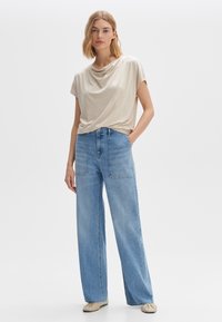 Top beige a maniche corte drappeggiato abbinato a jeans azzurri a vita alta e gamba larga. L'insieme presenta una vestibilità comoda e una texture morbida.