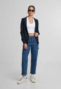 Felpa zip-up blu navy, canotta corta bianca, jeans blu a vita alta, sneakers bianche con suola crema. Gli accessori includono occhiali da sole neri.