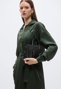 Πράσινη σατέν ολόσωμη φόρμα με φερμουάρ στο γιακά και τσέπες, κρατώντας μια μικρή μαύρη τσάντα με υφή και διπλές λαβές.