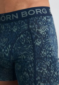 Tengerés és zöld mintázatú boxer nadrág, elasztikus anyagból készült, puha elasztikus derékkal, amelyen "BORG" felirat található.