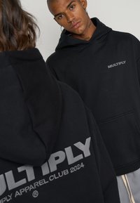 Black hooded sweatshirt featuring a front pocket and printed "MULTIPLY" logo on the chest and back, designed with a relaxed fit.