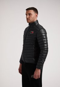 Giubbino imbottito nero con colletto alto, apertura con zip e logo ricamato sul petto. Finitura opaca con una texture trapuntata.