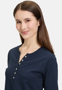 Granatowa bluzka z długim rękawem o zaokrąglonym dekolcie, z pięcioma metalowymi guzikami wzdłuż zapięcia. Wykonana z gładkiego materiału bawełnianego.