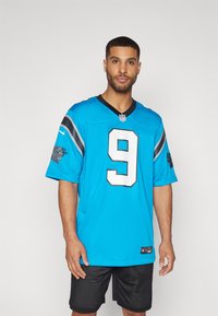 Kék NFL mez, amelyen a 9-es szám és a csapat logója található a mandzsettákon, fekete V-nyakú gallérral. Fekete hálós sorttal viselve.
