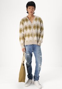 Cárdigan de lana beige con patrón de diamantes verdes, con botones en el frente y puños de canalé. Lleva puestos unos vaqueros azul desgastados y unas zapatillas blancas, sosteniendo un bolso en tono marrón claro.