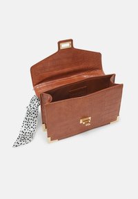 Pochette marron en simili cuir crocodile avec coins dorés, fermoir doré, rabat ouvert et tissu tacheté noir et blanc noué sur le côté.