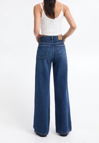 Džíny s vysokým pasem a širokými nohavicemi v tmavě modrém denimovém provedení, s klasickým pětiokamžitým designem a obdélníkovou zadní nášivkou.