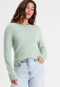 Mintgrøn sweater med et struktureret honningkubemønster, rund halsudskæring og ribbede manchetter. Båret med lyseblå denimjeans.