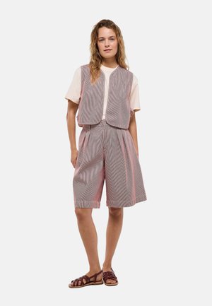Femme aux cheveux ondulés porte une chemise beige à manches courtes, un gilet gris sans manches, un short plissé assorti jusqu'aux genoux et des sandales plates marron.