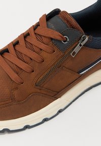 Brun avslappnad sneaker med strukturerad tygtunga, runda snörningar, sidoglid och vit gummisula med blå kant.