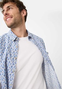 Korte mouwen bloeshemd in blauwe en beige tinten, met een button-down kraag en een lichte stof, gedragen over een wit t-shirt.
