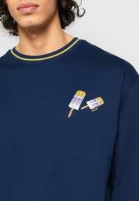 Sweatshirt bleu marine avec un col côtelé à rayures jaunes et vertes. Présente des esquimaux brodés en blanc, violet et jaune avec le détail "MCM".