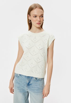 Witte gebreide trui met een hexagonaal patroon, korte mouwen en een textuurafwerking; gecombineerd met lichtblauwe denim jeans.