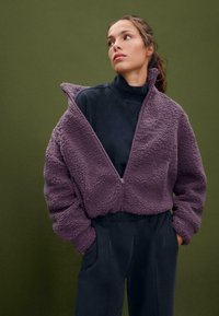 Giacca in pile viola con zip, caratterizzata da una texture morbida e una vestibilità oversize, indossata sopra un top nero a collo alto, abbinata a pantaloni scuri.