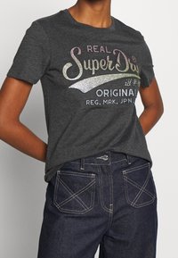 Grå bomulds t-shirt med glitrende, multikolor tekst, der siger "REAL SuperDry ORIGINAL", parret med mørk denim jeans med kontrastsyninger.