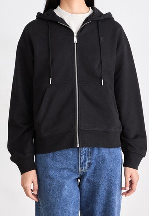 Personne portant un sweat à capuche noir avec fermeture éclair et poches avant, par-dessus une chemise blanche et un jean bleu, debout devant un fond clair uni.