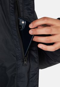 Giacca navy con una texture liscia e lucida; mano che inserisce uno smartphone in una tasca laterale con zip. Accentuata da un piccolo tirazip.