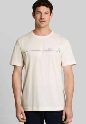 Mann trägt ein hellbeiges T-Shirt mit kurzen Ärmeln mit einem kleinen blauen Segelboot-Grafik und Text auf der Brust, steht vor einem einfarbigen Hintergrund.