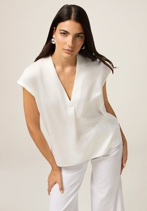 White, short-sleeved blouse with a deep V-neck and soft, smooth fabric, paired with white tailored pants. Long, straight hair and large earrings.