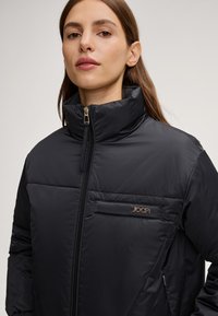 Schwarze gepolsterte Jacke mit hohem Kragen, Reißverschlussschluss und zwei Reißverschlusstaschen. Mit einem kleinen metallischen Logo auf der Brust. Glatte Textur.