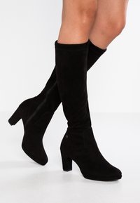 Botas altas de muslo negras de ante, con puntera redondeada, tacón cuadrado y cremallera lateral para facilitar su uso. Textura suave con un ligero brillo.