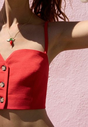 Rotes, kurzes Top mit dünnen Trägern, strukturiertem Stoff und drei dekorativen Knöpfen in der Mitte, kombiniert mit einer Chilipfeffer-Anhänger-Halskette.