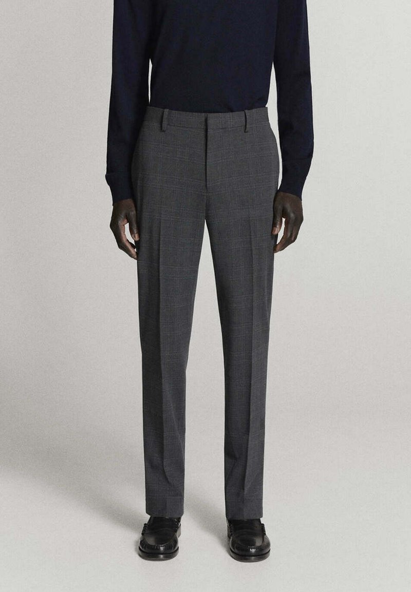 Homme portant un pantalon de costume gris à carreaux, une chemise noire à manches longues et des chaussures noires, debout sur un fond uni.