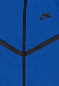 Modrá zapínacia bunda vyrobená z hladkej tkaniny. Obsahuje čierne lemovanie a čierne logo Nike na pravej strane.
