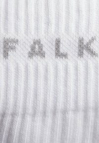 Calza bianca a coste con testo grigio che legge "FALK." Il tessuto è morbido e testurizzato, con motivi a maglia visibili.