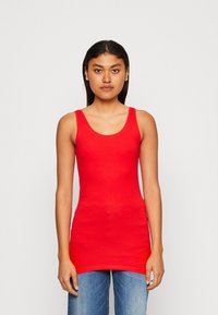 Top sin mangas de algodón rojo con escote redondeado y corte ajustado, combinado con pantalones vaqueros de mezclilla azul. La tela parece suave y sin adornos.