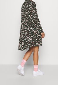 Femme portant une robe florale vert foncé, des chaussettes rayées roses et des baskets blanches, se tenant avec un pied légèrement levé contre un fond uni.