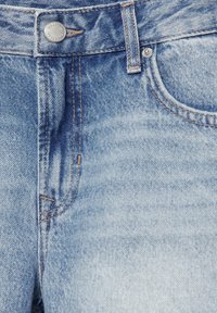 Gros plan sur un jean en denim bleu clair montrant un bouton, une boucle de ceinture, des coutures et une poche avant avec rivet en métal.