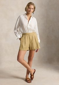 Γυναίκα με άσπρο πουκάμισο με κουμπιά, μπεζ σορτς ψηλής μέσης, καφέ σανδάλια και πολύχρωμα σκουλαρίκια με κρεμαστά στοιχεία, στέκεται με το ένα πόδι σταυρωμένο.